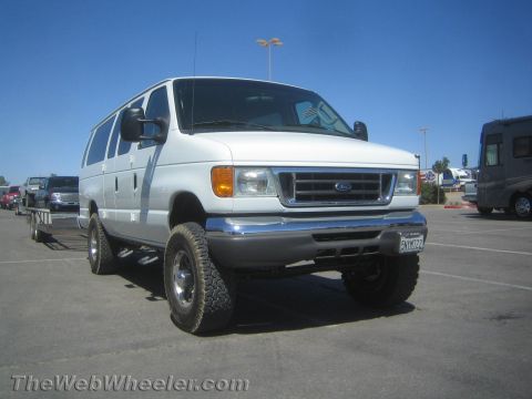 1990 Ford Bronco - Front three-quarter view of a white lifted Ford E-series passenger van on off-road tires parked beside a motorhome.