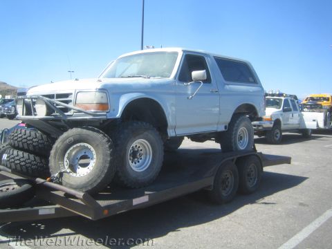 1990 Ford Bronco - Light blue full-size Ford Bronco on off-road tires loaded on a flatbed trailer with spare wheels and desert hills behind.