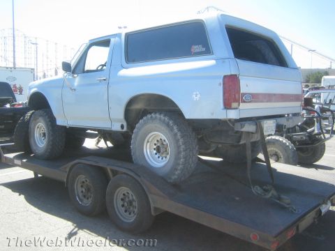 1990 Ford Bronco - light blue full-size Ford Bronco with off-road tires sits strapped down on a tandem-axle flatbed trailer in a paved lot.