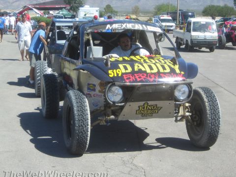 1990 Ford Bronco - man drives a single-seat off-road race buggy with Mexicali and yellow energy-drink lettering through a paved staging area.