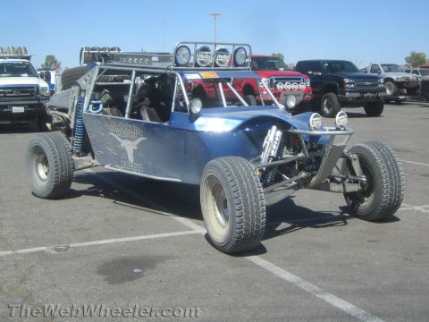 1990 Ford Bronco - blue four-seat off-road sand rail buggy with light bar and long-travel suspension parked among trucks in a paved lot.