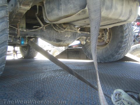 1990 Ford Bronco - Underbody view of a vehicle on a trailer showing the chassis, exhaust, oversized tire and tensioned ratchet straps on a diamond-plate deck.