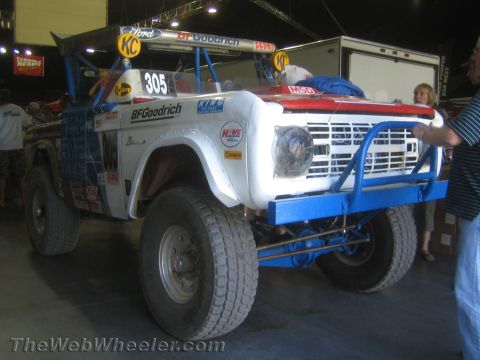 1990 Ford Bronco - Side-front view of a white early Ford Bronco race truck, number 305, with BFGoodrich graphics, blue tube work and large off-road tires.