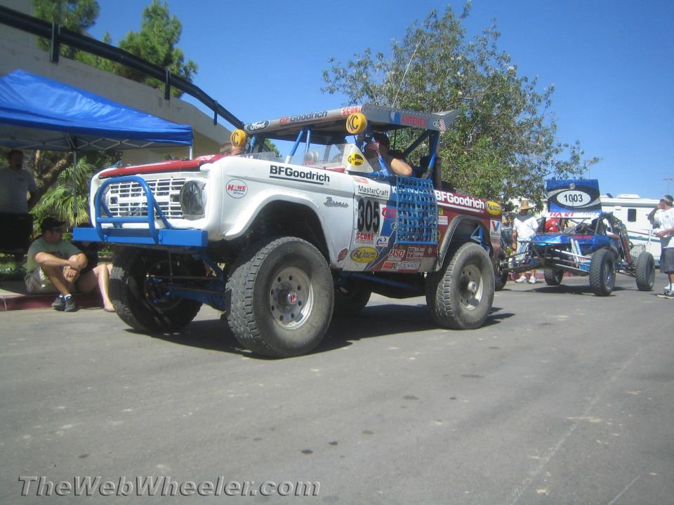 White Ford Bronco race truck no. 805 with blue bumper parked outdoors near tents and a second buggy