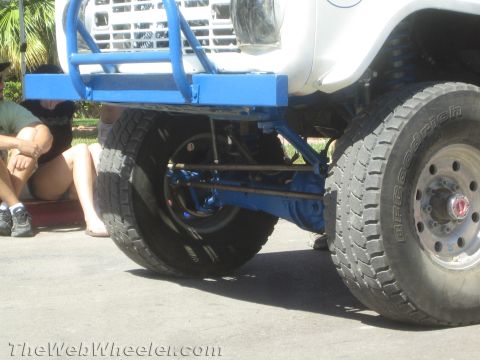 1990 Ford Bronco - Low-angle view under the front of a white off-road race truck showing the blue tube bumper, axle and BFGoodrich tires.