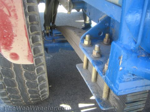 1990 Ford Bronco - Detail of a blue frame bracket with large bolts, a leaf-spring stack and an off-road tire on a race truck.