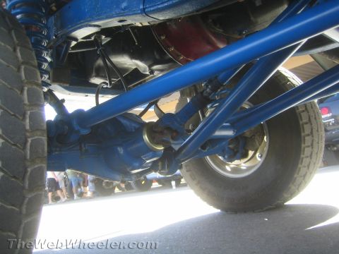 1990 Ford Bronco - Underbody view of an off-road race truck showing the blue-painted rear axle, suspension links, driveshaft and tire.