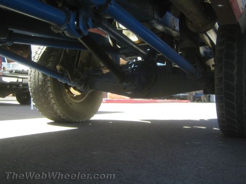 1990 Ford Bronco - Backlit low-angle view under an off-road race truck showing the blue axle housing, links and tire over asphalt.