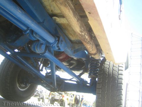1990 Ford Bronco - Underbody view of an off-road race truck with driveshaft, blue suspension links, a coil shock and a wood-floored bed above.