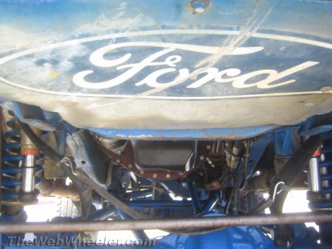1990 Ford Bronco - Underbody view of a blue fuel tank bearing the Ford oval logo mounted above the rear axle and leaf springs of an off-road race truck.