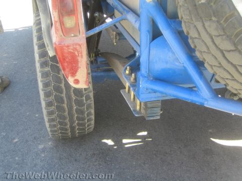 1990 Ford Bronco - Side detail of the blue tube frame, leaf-spring stack and tires beneath a red-fendered off-road race truck on asphalt.