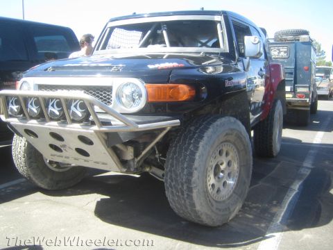 1990 Ford Bronco - Front three-quarter view of a black Toyota FJ Cruiser off-road race truck with KC driving lights, tube bumper and beadlock wheels in a lot.