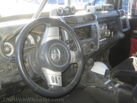 1990 Ford Bronco - Interior of a Toyota-badged off-road race truck showing the steering wheel, dash gauges, shifter and roll cage padding.