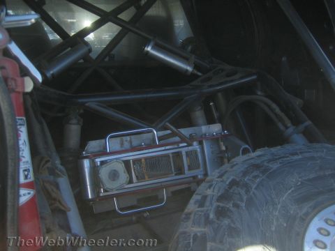 1990 Ford Bronco - Dim view inside the rear roll cage of an off-road race truck showing a mounted device, bypass shocks and an off-road tire.
