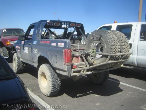 1990 Ford Bronco - blue lifted Ford-style off-road prerunner numbered 349 with rear-mounted spare tires and a tubular bumper sits in a paved lot.