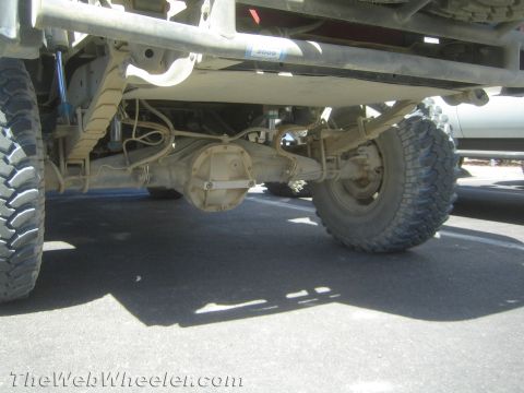 1990 Ford Bronco - underside of a lifted off-road truck reveals a solid axle, leaf springs, exhaust piping and oversized mud tires.