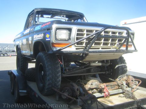 1990 Ford Bronco - black-and-silver vintage Ford 4x4 truck with a tube grille guard sits chained to a flatbed trailer beside loose drivetrain parts.