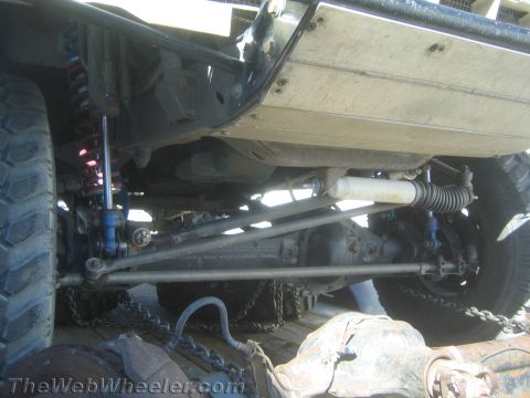 1990 Ford Bronco - undercarriage of a lifted truck on a trailer reveals the front axle, coilover shocks, steering linkage and tie-down chains.