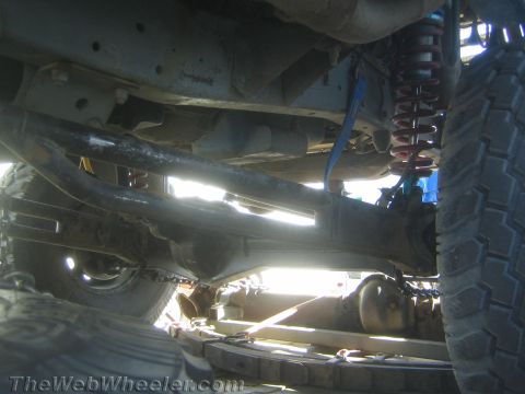 1990 Ford Bronco - wide view beneath a lifted truck shows the axle, driveshaft, a red-and-blue coilover and large tires backlit by sunlight.