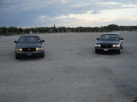 2000 Ford Crown Victoria - Two Ford Crown Victorias are parked in a large, empty lot under a cloudy sky.