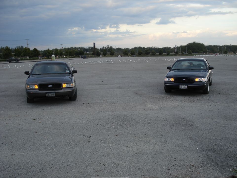 Two dark Ford Crown Victoria sedans facing camera in an empty lot under cloudy sky