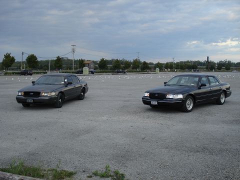 2000 Ford Crown Victoria - Two Ford Crown Victoria police cars are parked in an open lot with a cloudy sky overhead.