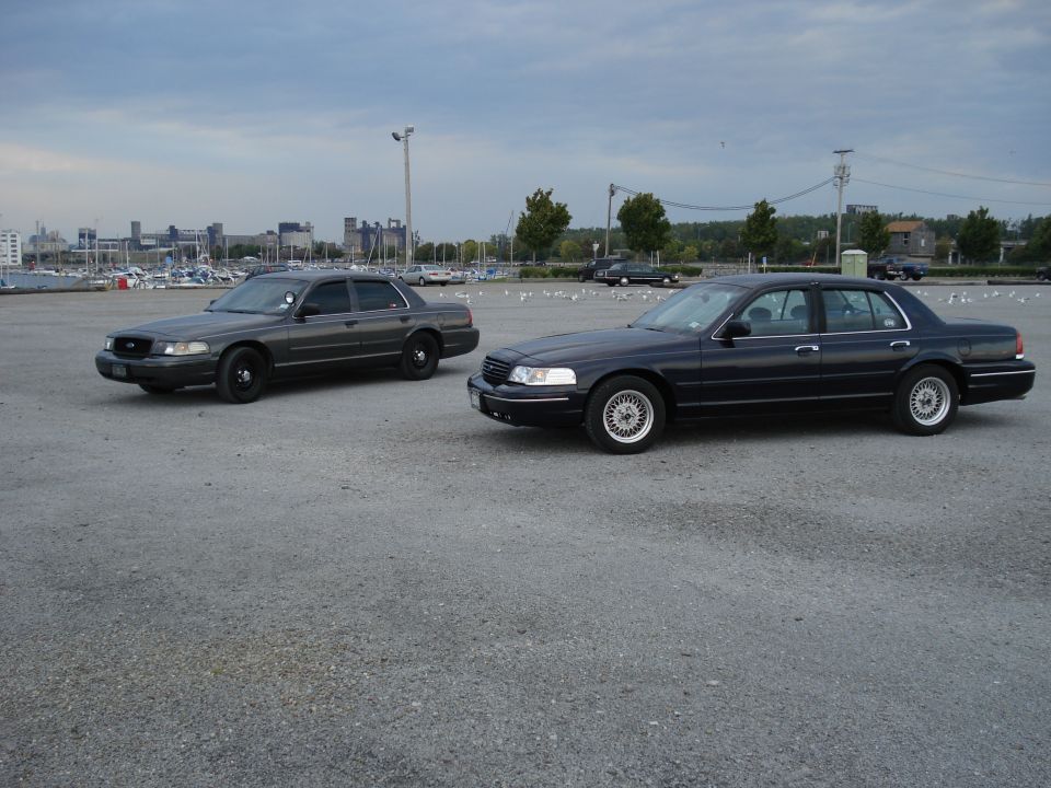 Gray police-spec and dark blue Ford Crown Victoria sedans in a gravel lot by a marina
