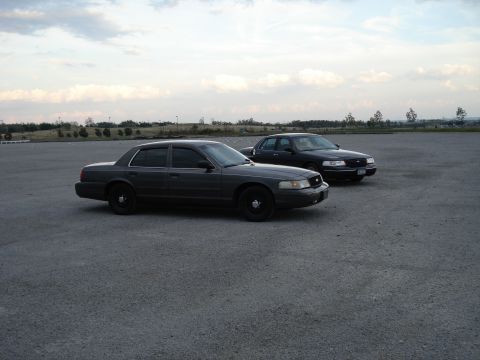 2000 Ford Crown Victoria - Two Ford Crown Victorias are parked in an open lot under a cloudy sky.