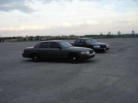 2000 Ford Crown Victoria - Two Ford Crown Victorias are parked in an open lot under a cloudy sky.