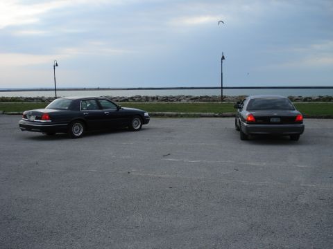 2000 Ford Crown Victoria - Two Ford Crown Victorias are parked in a lot next to a body of water under a cloudy sky.