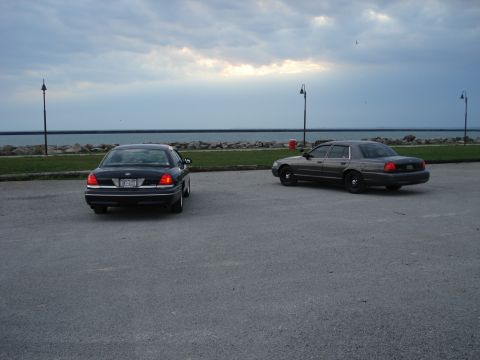 2000 Ford Crown Victoria - Two Ford Crown Victoria sedans are parked on asphalt near a rocky shoreline under a cloudy sky.