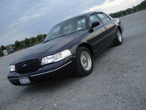 2000 Ford Crown Victoria - A dark blue 2000 Ford Crown Victoria is parked on a gravel surface under a cloudy sky.