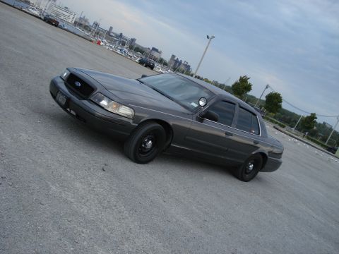2000 Ford Crown Victoria - A dark grey 2000 Ford Crown Victoria sedan is parked on a gravel lot near a marina.