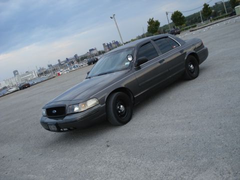 2000 Ford Crown Victoria - Dark gray Ford Crown Victoria police car parked on asphalt near a marina with city skyline in background.