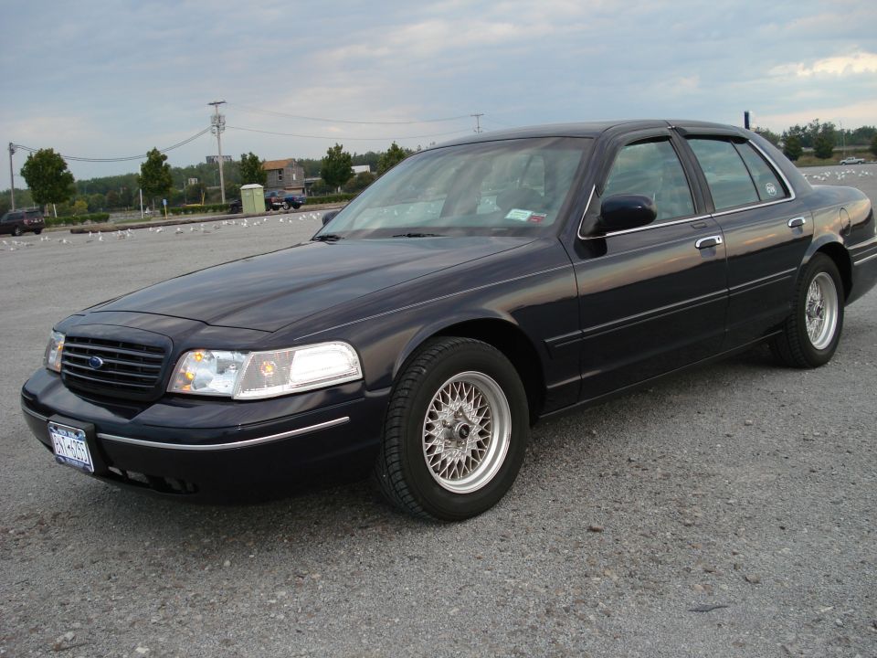 Dark blue Ford Crown Victoria sedan with wire-spoke wheels parked on a gravel waterfront lot