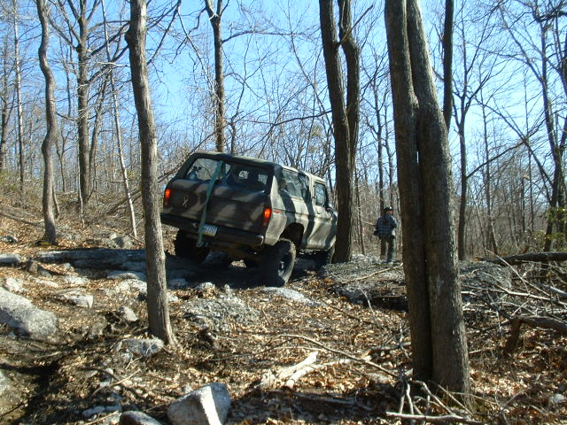 Camouflaged SUV navigating a rocky, wooded trail with a person standing nearby