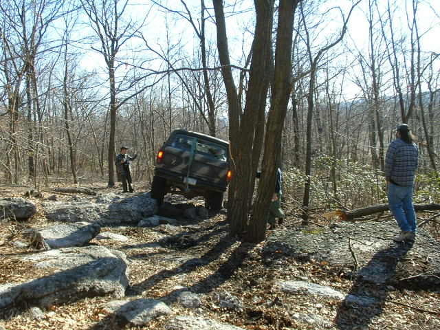 SUV driving over rocks in a wooded area. Two people are watching