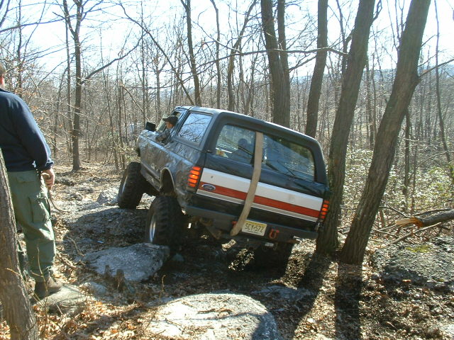 Ford Bronco with lifted suspension navigating rocks on a wooded trail. The rear of the vehicle shows a white stripe with a red accent