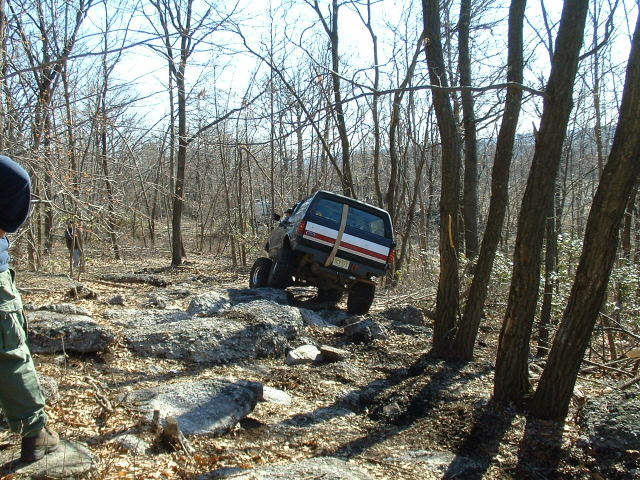 SUV with red and white stripes on rear, going over rocks in a wooded area