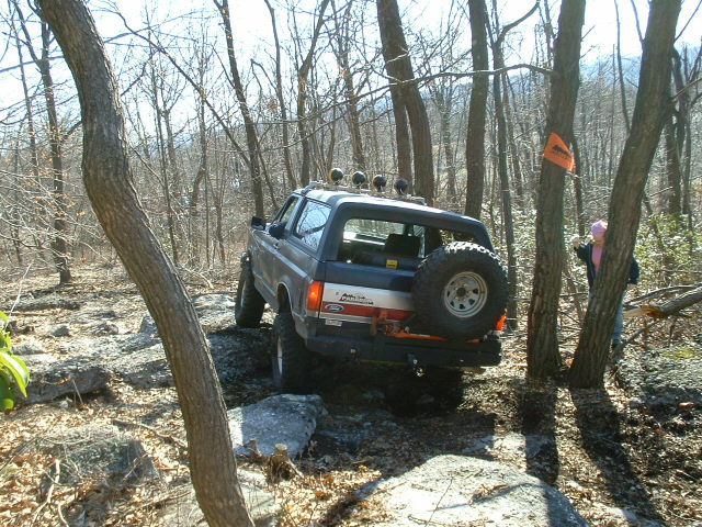 Ford Bronco off-roading on a rocky, wooded trail. Spare tire mounted on the back