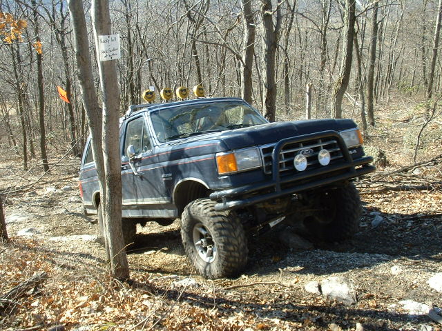 Dark blue Ford Bronco with off-road tires and lights on roof driving on a wooded trail