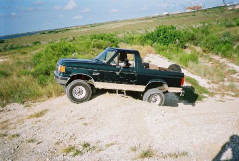 1990 Ford Bronco - This Ford Bronco is navigating a sandy, off-road terrain with one wheel partially buried.