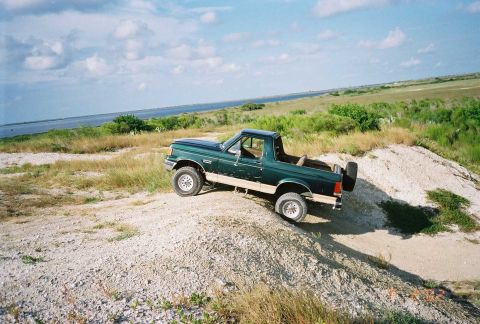 1990 Ford Bronco - A green Ford Bronco navigates a dirt incline near a body of water under a cloudy sky.