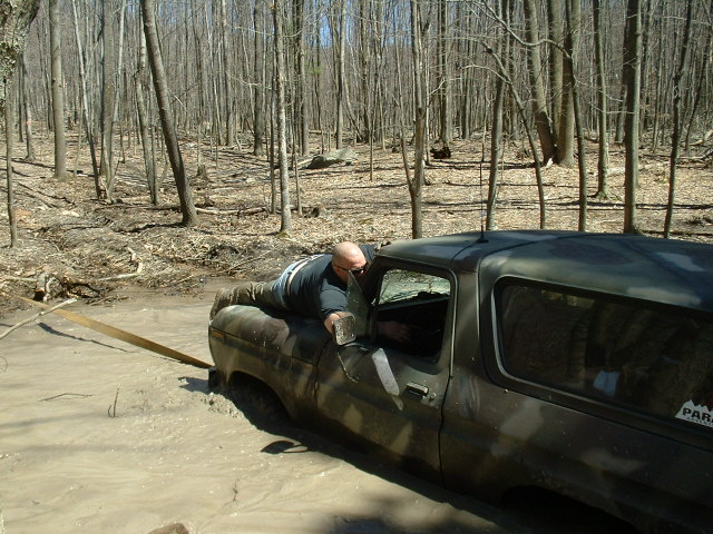 Man lying on side of dark off-road vehicle stuck in mud, trees in background