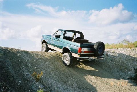 1990 Ford Bronco - This 1990 Ford Bronco is captured in a three-quarter front view as it navigates an incline on a dirt path under a cloudy sky.