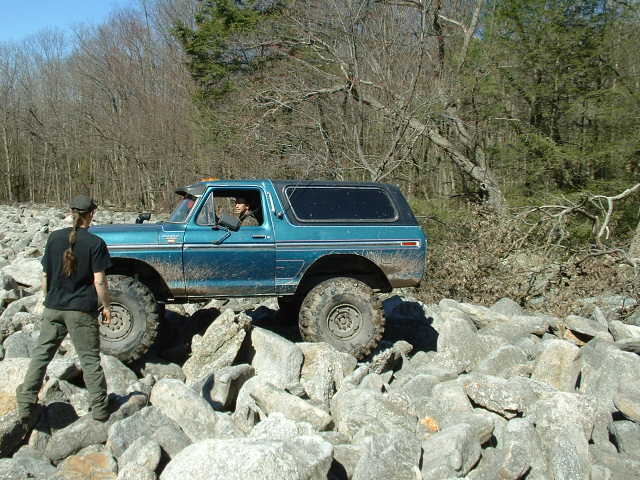 Blue 1978 Ford Bronco off-roading over large rocks, with a person standing nearby