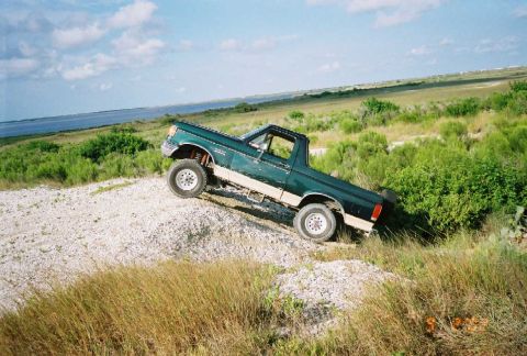 1990 Ford Bronco - 1990 Ford Bronco navigating an off-road incline with a scenic backdrop of water and vegetation.