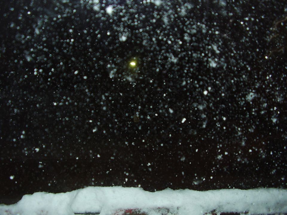 Snow falling at night with a yellow light in the distance and snow on the ground