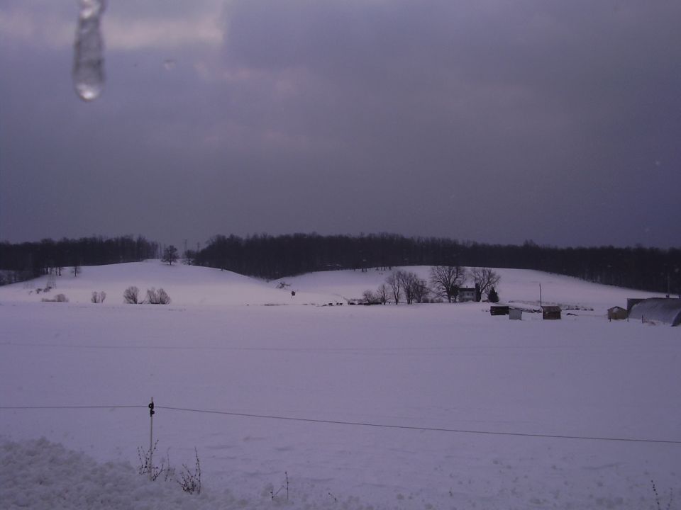 Snowy field with scattered buildings and trees under a cloudy sky. A water droplet hangs in the upper left