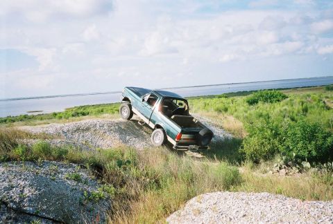1990 Ford Bronco - This truck is on a dirt path with a body of water in the background under a cloudy sky.